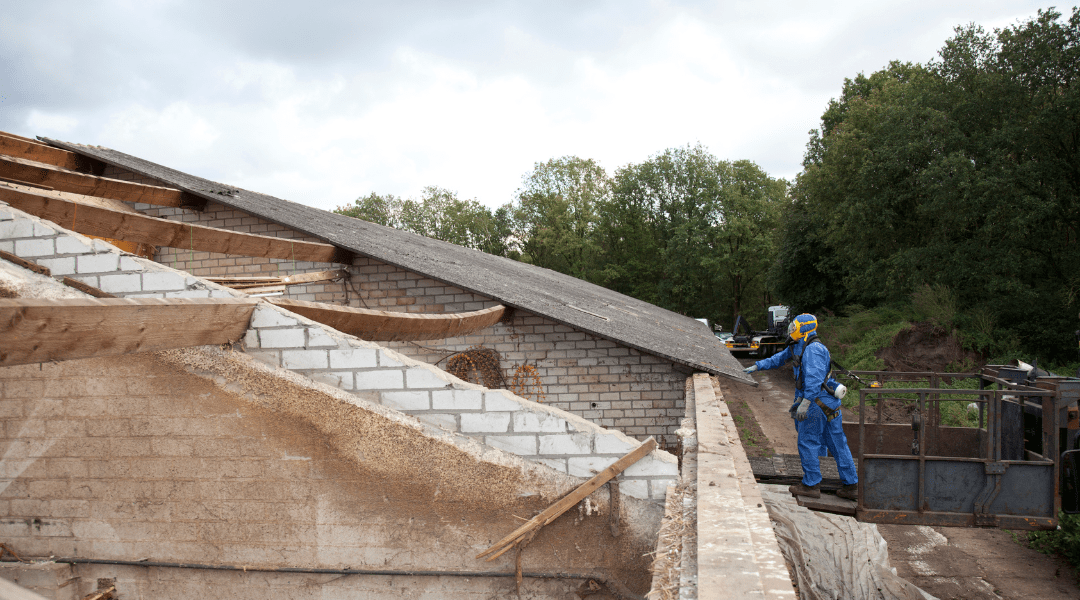 Sicheres Entfernen von Asbestmaterialien auf einem Dach Ein Arbeiter in Schutzkleidung führt eine Asbestsanierung auf einem teilweise abgedeckten Dach eines Gebäudes durch.